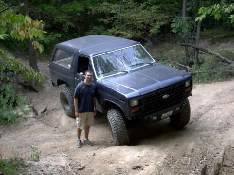 1986 Ford Bronco - A man stands beside a lifted Ford Bronco as it navigates a dirt path through a wooded area.