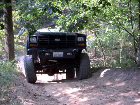 1986 Ford Bronco - A lifted black Ford Bronco navigates a wooded trail from a low front-facing angle.