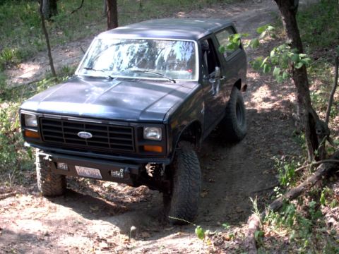 1986 Ford Bronco - This 1986 Ford Bronco is captured from a 3q-front perspective, navigating a wooded dirt path with an aftermarket bumper.