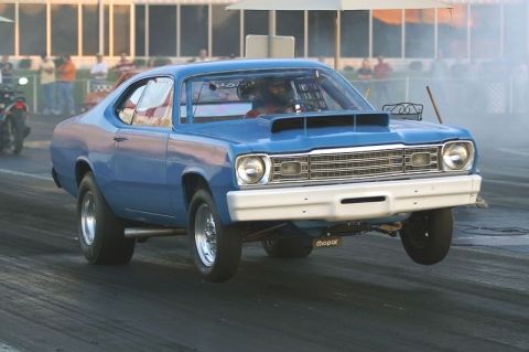 1989 Ford Bronco - A blue Mopar drag car lifts its front wheels off the ground during a race, kicking up smoke.