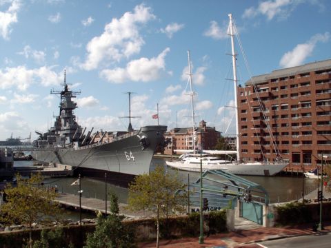 1988 Ford Bronco - A large battleship and a modern yacht are moored in a harbor with a brick building in the background.
