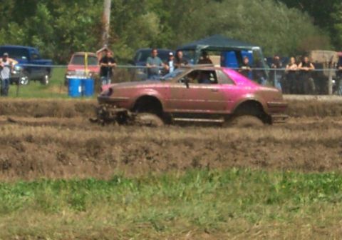 1979 Ford F-150 - A pink coupe navigates a mud pit during an event, with spectators in the background.