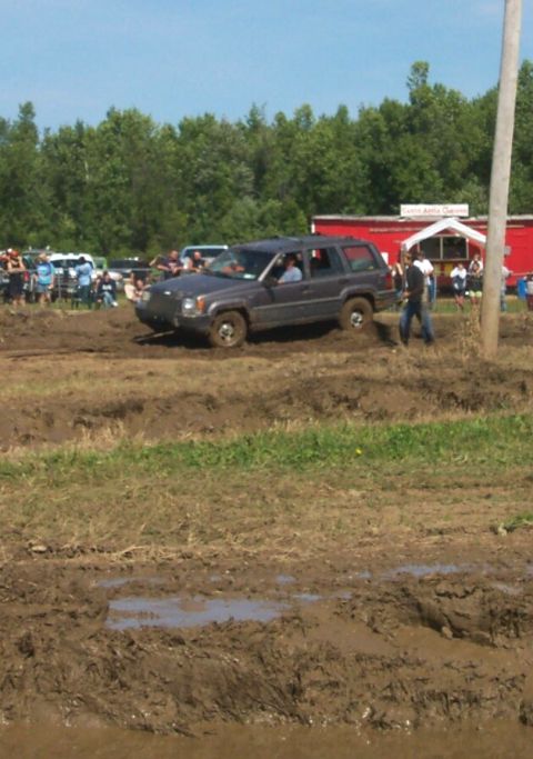 1979 Ford F-150 - A gray SUV is seen driving through a muddy obstacle course with onlookers and a building visible in the distance.