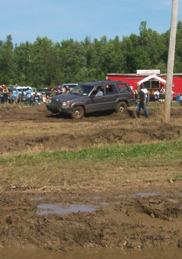 Gray SUV navigating a muddy track with spectators and a red building in the background