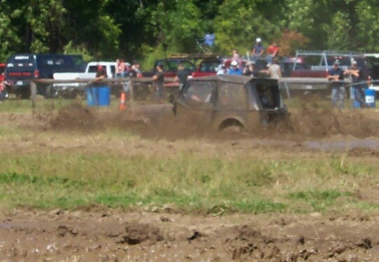 Muddy off-road vehicle driving through a mud pit with spectators in the background