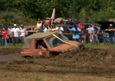 1979 Ford F-150 - A red off-road vehicle is stuck in a mud pit during an event, with a crowd of people watching from the sidelines.