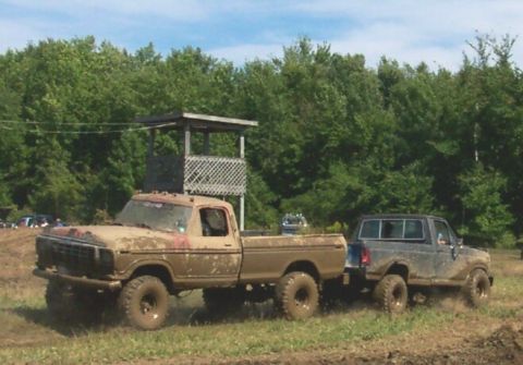 1979 Ford F-150 - Two trucks covered in mud are in an outdoor area with trees and a wooden structure.