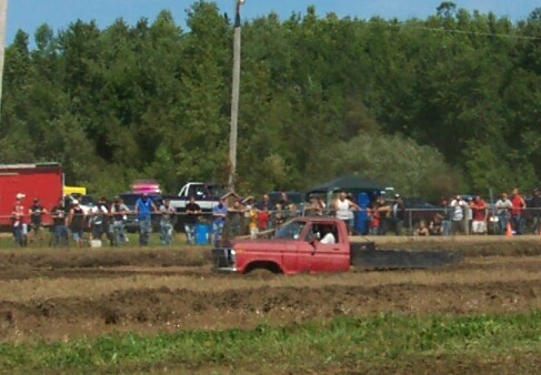 Red pickup truck stuck in mud, side view. Visible grille and front fender. Crowd watches in background