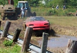 1979 Ford F-150 - A red sports car navigates a muddy track, with tractors visible in the background during an event.