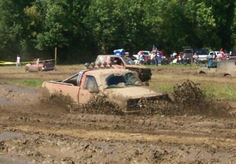1979 Ford F-150 - Several trucks are navigating a muddy track during an event, with trees and spectators visible.