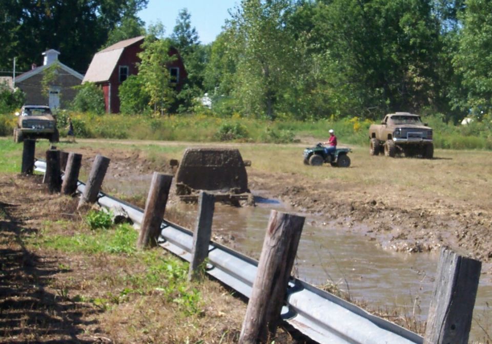 Trucks stuck in mud, one on posts, another in a pit, and an ATV driving nearby