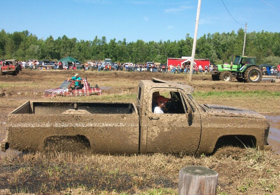Muddy pickup truck stuck in a mud pit at an event. Another truck and a tractor are visible