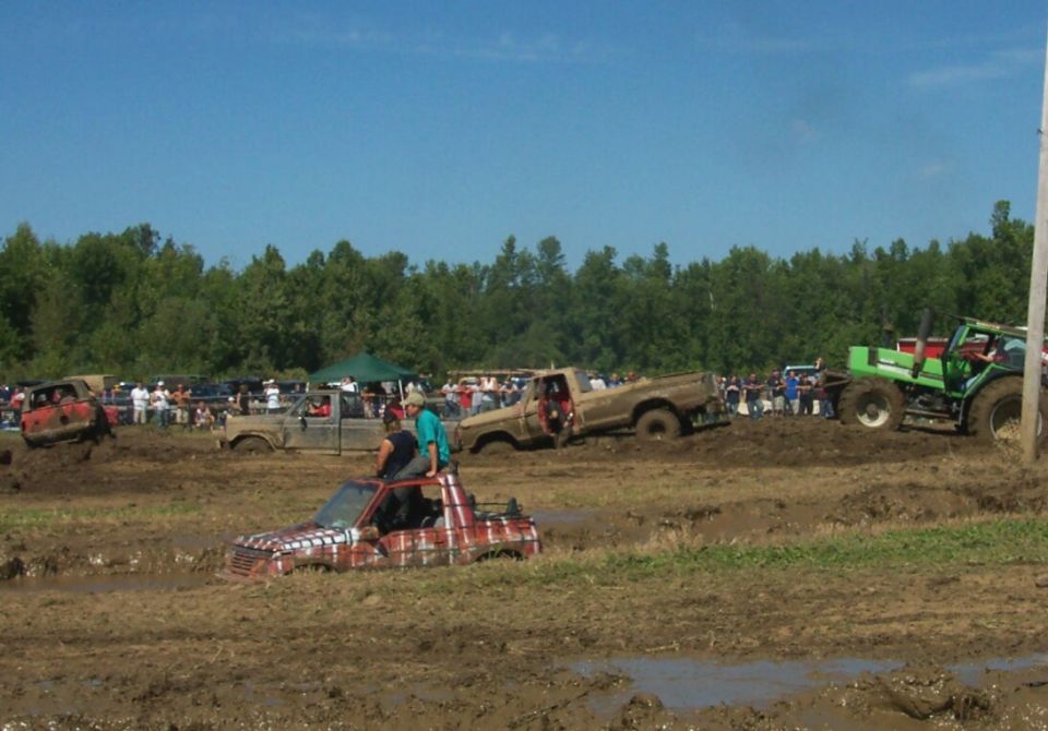 Mud bog event with several trucks stuck in the mud and a green tractor pulling one