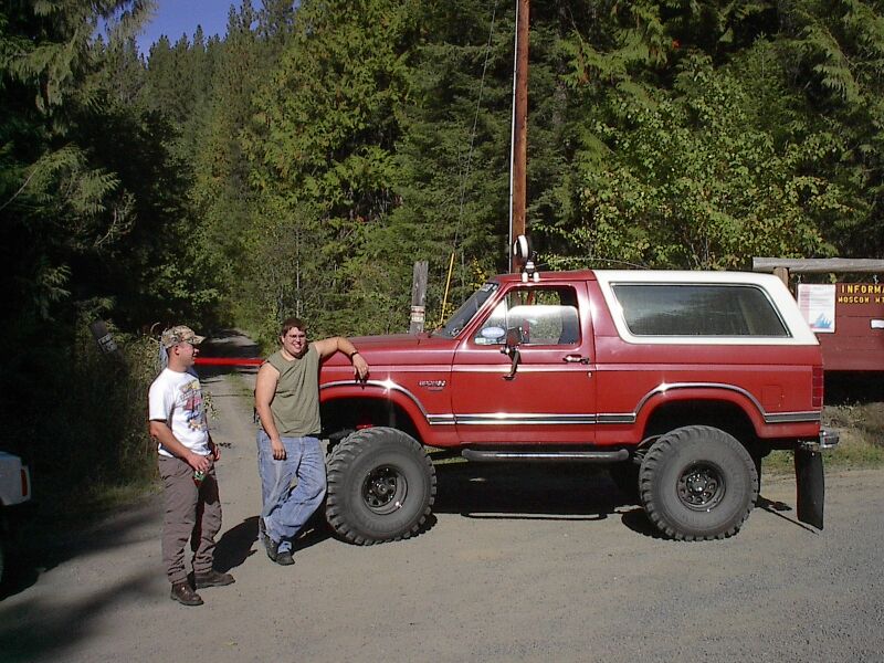 Red lifted two-door Ford Bronco with white top on gravel road, two men standing beside it