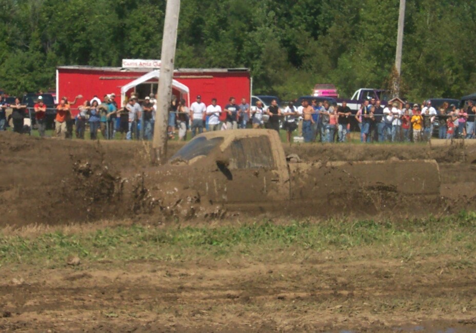 Muddy truck partially submerged in a mud pit with spectators in the background