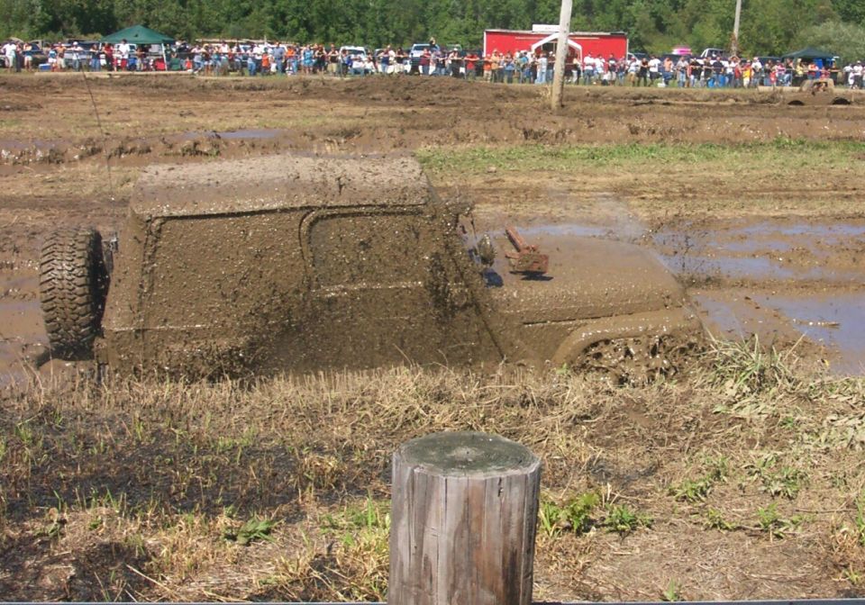 Mud-covered off-road vehicle stuck in a mud pit, side view. A spare tire is visible on the left