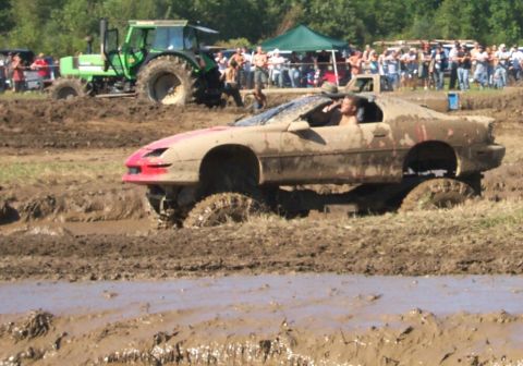 1979 Ford F-150 - A car navigates a muddy obstacle course during a competition, with a large crowd observing the event.