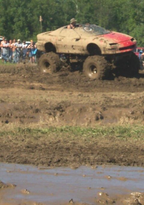 1979 Ford F-150 - A modified car body sits atop a monster truck chassis, navigating a challenging mud bog with spectators in the background.