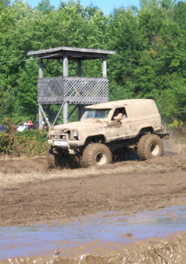 Mud-covered off-road vehicle with large tires navigating a muddy track near a wooden observation tower