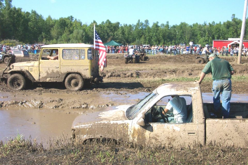 Mud bogging event with a yellow SUV and a pickup truck stuck in mud. Spectators watch from the background
