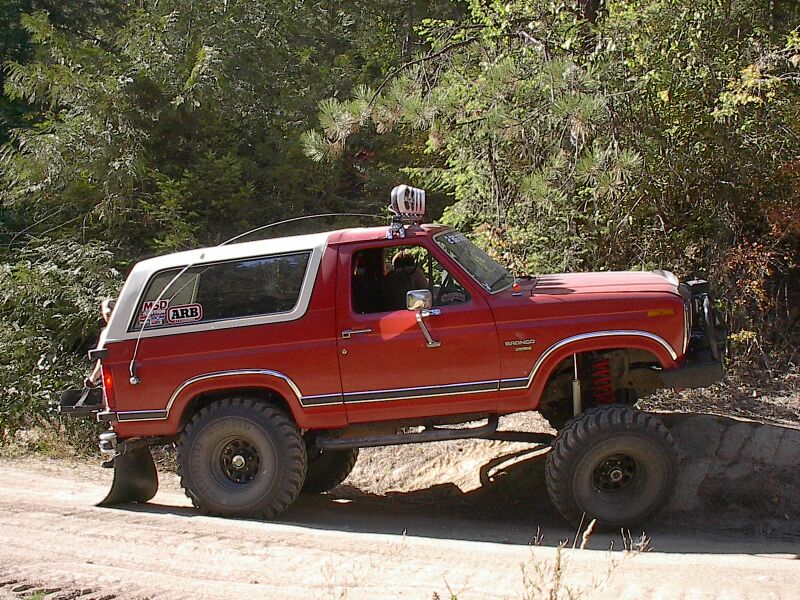 Lifted red and white Ford Bronco on off-road tires climbing a dirt embankment on a forest trail