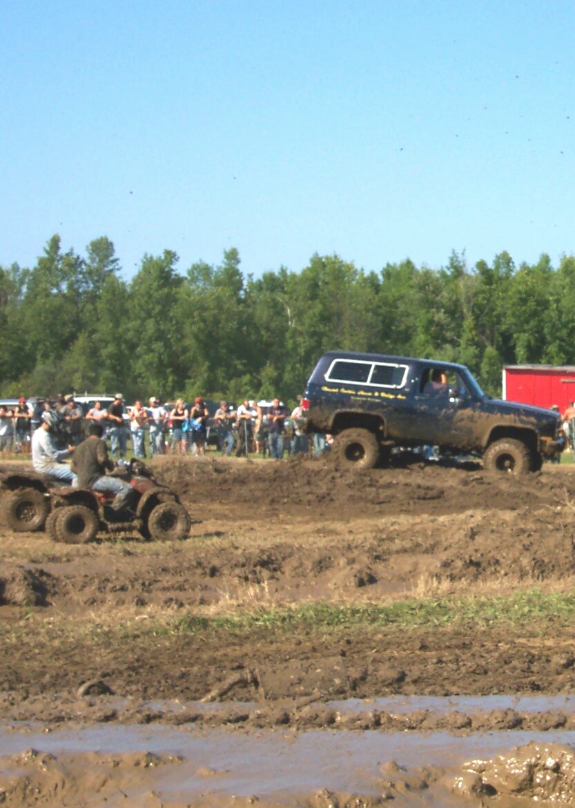 Muddy SUV and ATVs at a mud bogging event with a crowd watching