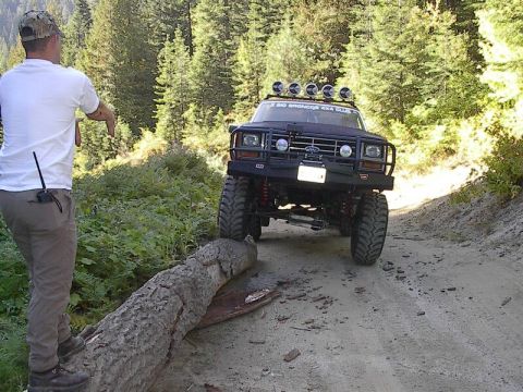 Lifted black Ford Bronco on a dirt trail straddling a fallen log, man in white shirt watching