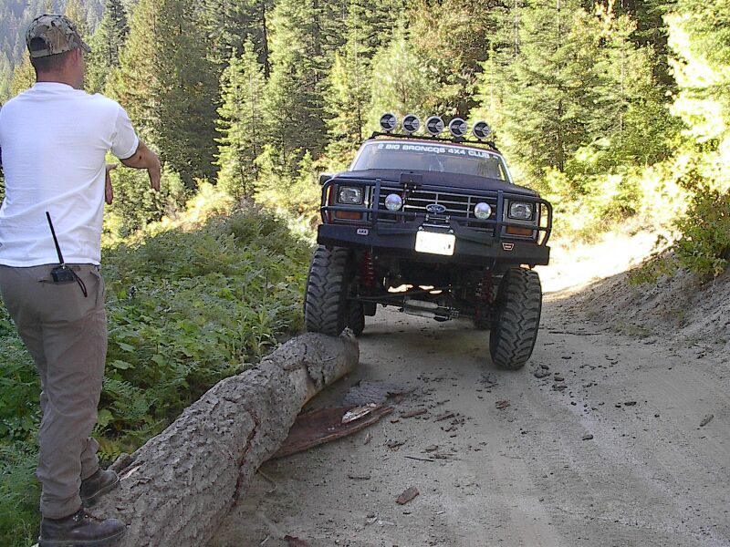 Lifted black Ford Bronco on a dirt trail straddling a fallen log, man in white shirt watching