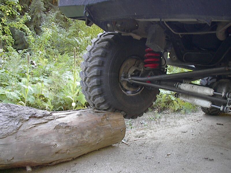 Lifted Bronco front tire climbing a log, showing red coil spring, dual shocks, and off-road tire