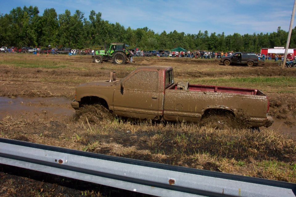 Mud-covered pickup truck stuck in mud pit with tractor and other vehicles in background