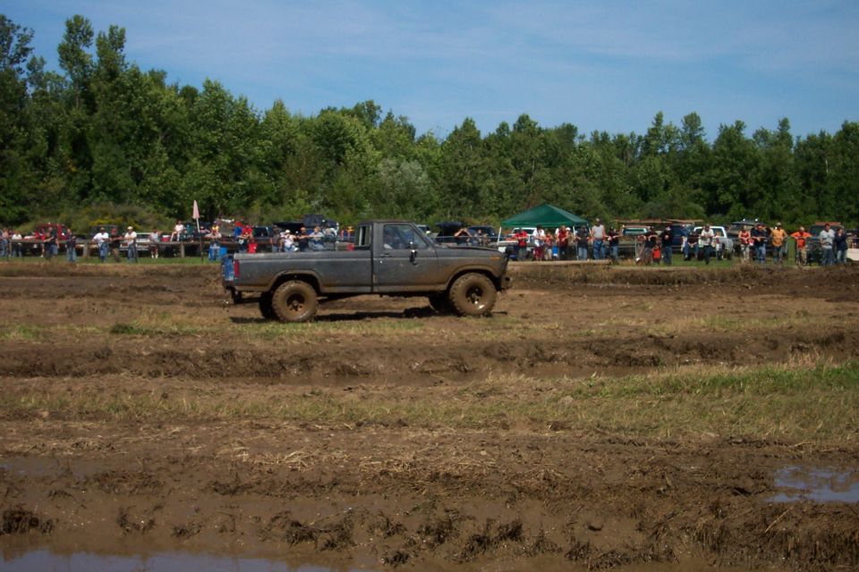 Gray pickup truck with large tires driving through mud, crowd and trees in background