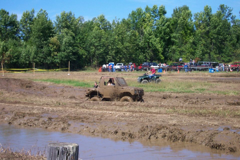 Mud-covered off-road vehicle stuck in a muddy field with an ATV nearby and spectators in the background
