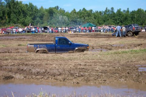 1979 Ford F-150 - A blue pickup truck is deeply stuck in a mud pit during an event, with a crowd watching from the sidelines.