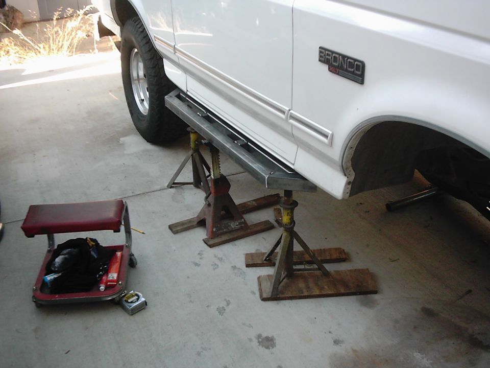 White Ford Bronco on jack stands with custom side steps, showing the rear fender and wheel well