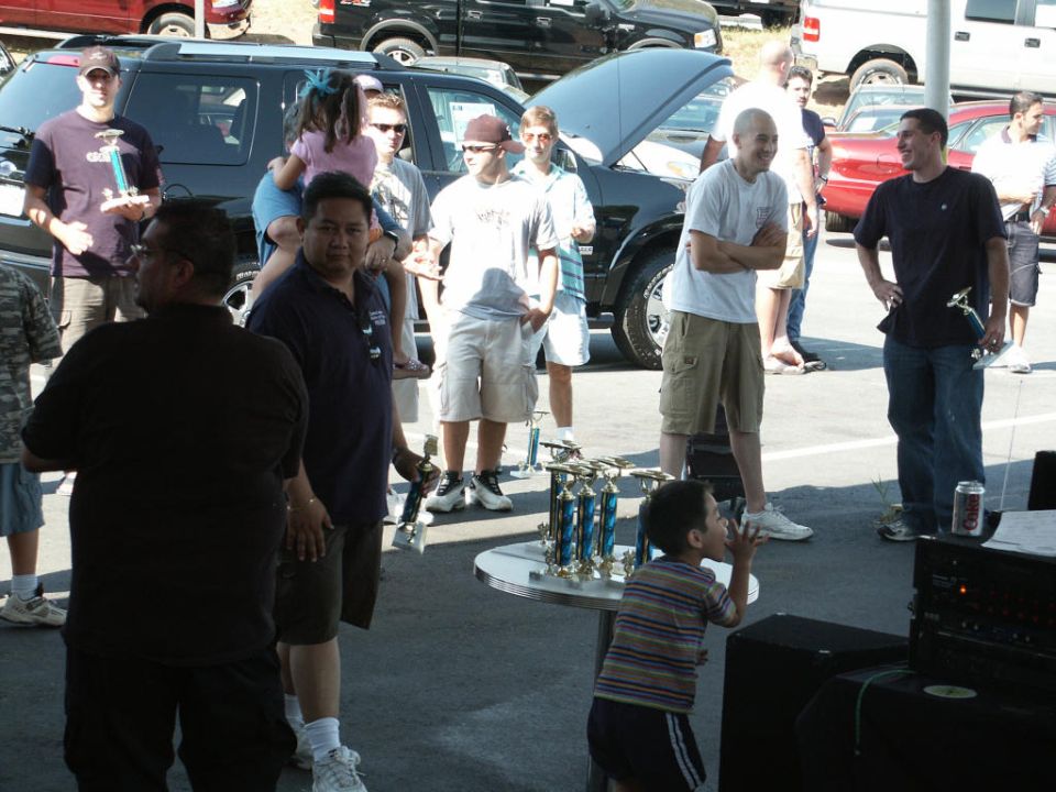 People gathered at an outdoor event with trophies on a table and several vehicles in the background