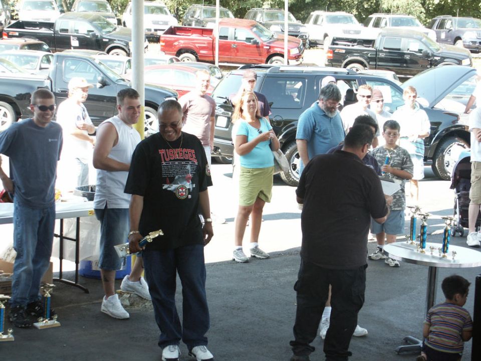 People gathered around tables with trophies at an outdoor event with many vehicles parked in the background