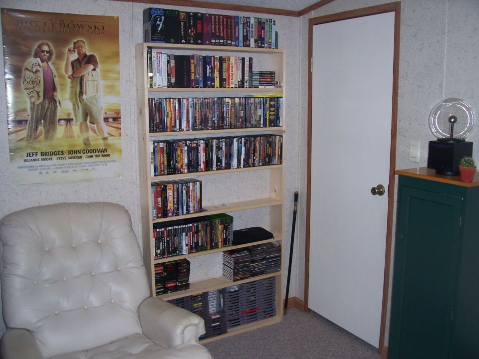 Room interior with a white leather chair, a poster for The Big Lebowski, and a tall bookshelf filled with DVDs and video games