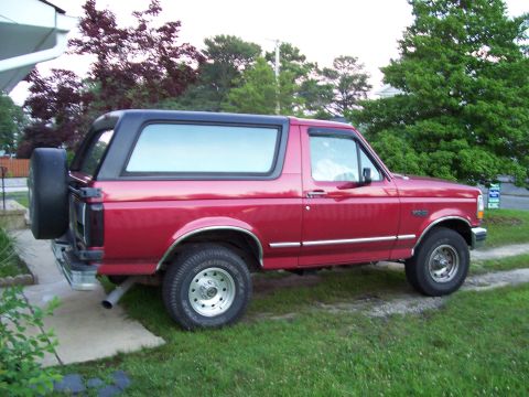 1995 Ford Bronco - This red Ford Bronco, likely from the early to mid-1990s, is shown from the side with its black top and rear-mounted spare tire.