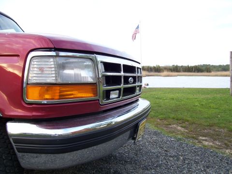 1995 Ford Bronco - The front grille and headlight assembly of a red Ford truck is shown parked near a body of water.