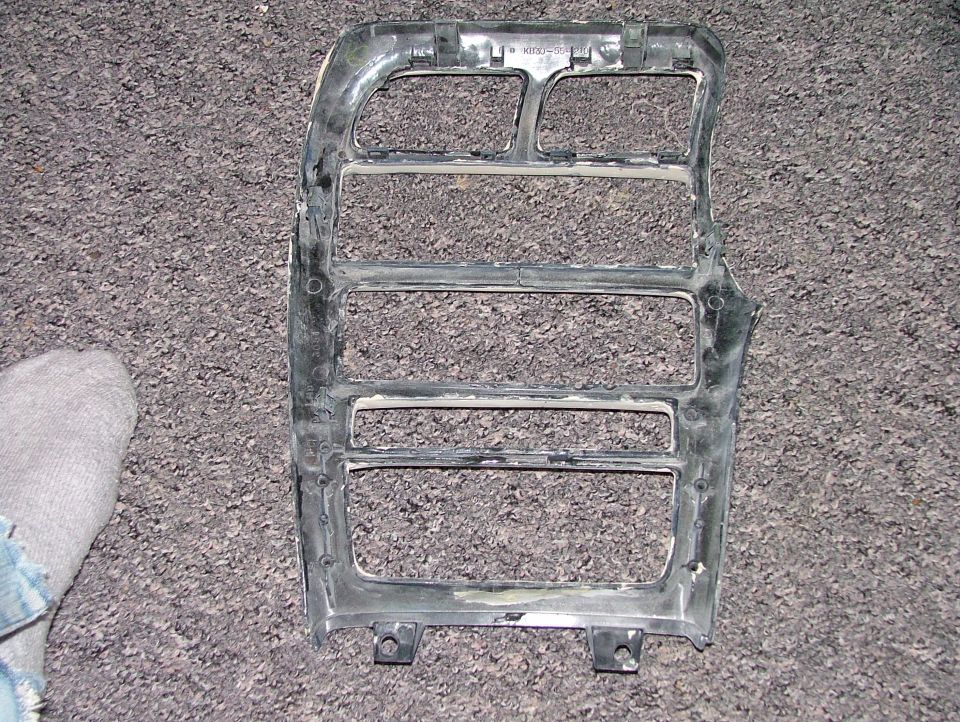 Close-up of a car's plastic grille assembly, possibly for a headlight area, lying on a carpeted surface. A person's socked foot is visible on the left