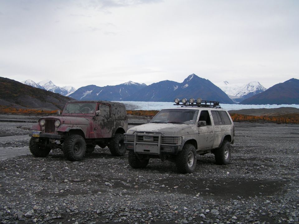 Muddy white lifted Nissan Pathfinder with roof lights beside red Jeep on gravel bar near glacier and mountains.