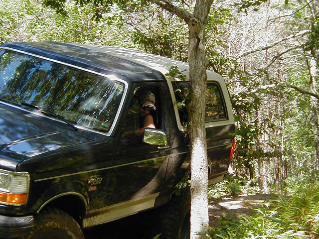 Black 1993 Ford Bronco with a white top, driving off-road near a tree