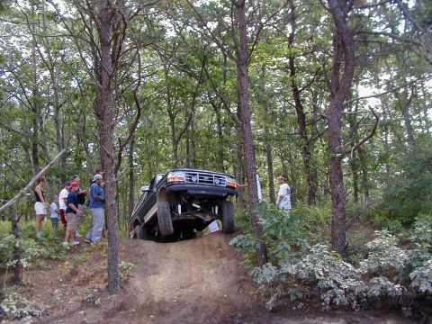 1993 Ford Bronco - A lifted dark green Ford Bronco crests a steep sandy hill on a wooded trail, front end pitched up so the front axle, shocks and skid pan ar…