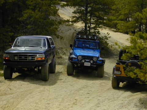 1993 Ford Bronco - Three off-road vehicles, including a 1993 Ford Bronco, are parked on a dirt path in a wooded area.