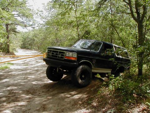 1993 Ford Bronco - Three-quarter front view of a lifted dark green Ford Bronco with tan lower stripe parked at the edge of a sandy two-track trail, front tire…