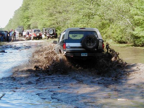 1993 Ford Bronco - Multiple off-road vehicles navigate a muddy trail with spectators watching from the side.