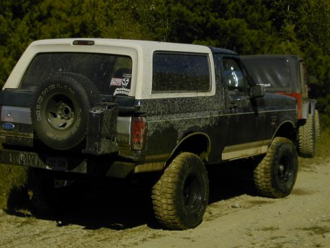 1993 Ford Bronco - Three-quarter rear view of a mud-splattered dark green Ford Bronco with a white removable hardtop on a sandy trail, wearing tall BFGoodrich…