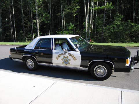 1991 Ford LTD Crown Victoria - Profile of a black-and-white Ford police sedan marked HIGHWAY PATROL with CHP star, parked on a road beside a pine forest.