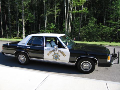 1991 Ford LTD Crown Victoria - Profile of a black-and-white Ford police sedan with HIGHWAY PATROL lettering and gold star emblem, parked on a road near woods.