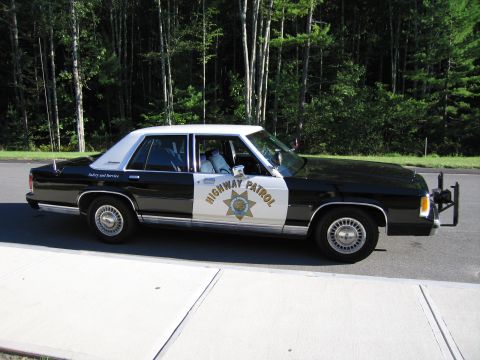 1991 Ford LTD Crown Victoria - Side profile of a black-and-white Ford police sedan marked HIGHWAY PATROL and Safety and Service, parked along a forested road.